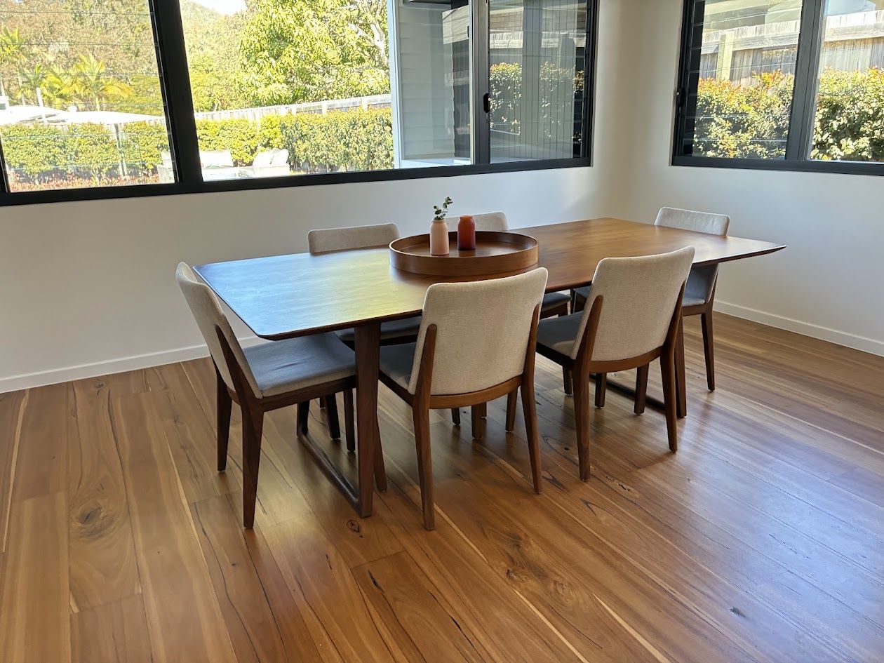 Timber dining table and upholstered chairs on wide hardwood boards beside large garden windows